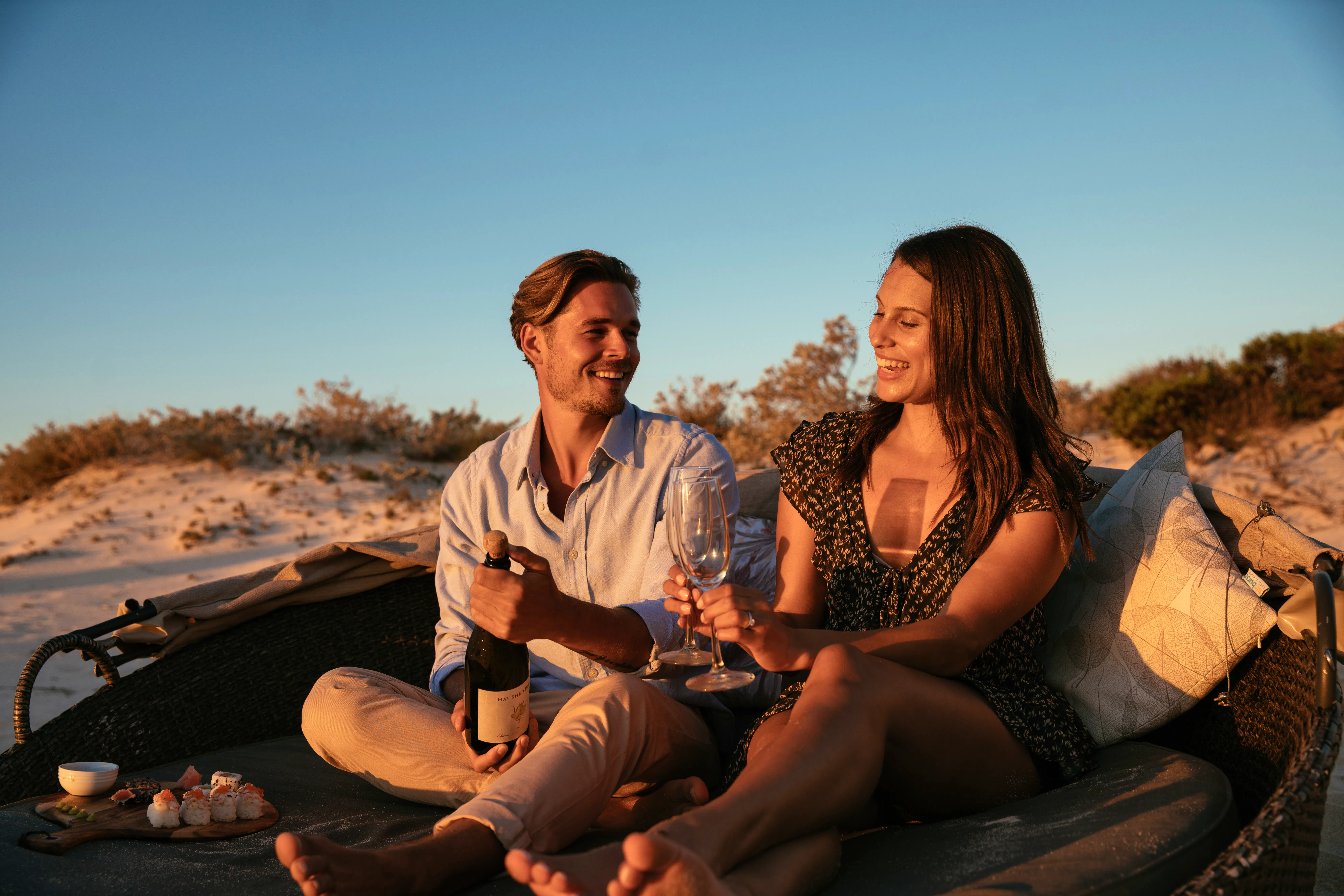 Smiling couple sharing champagne and sushi on a beach daybed at sunset at Sal Salis