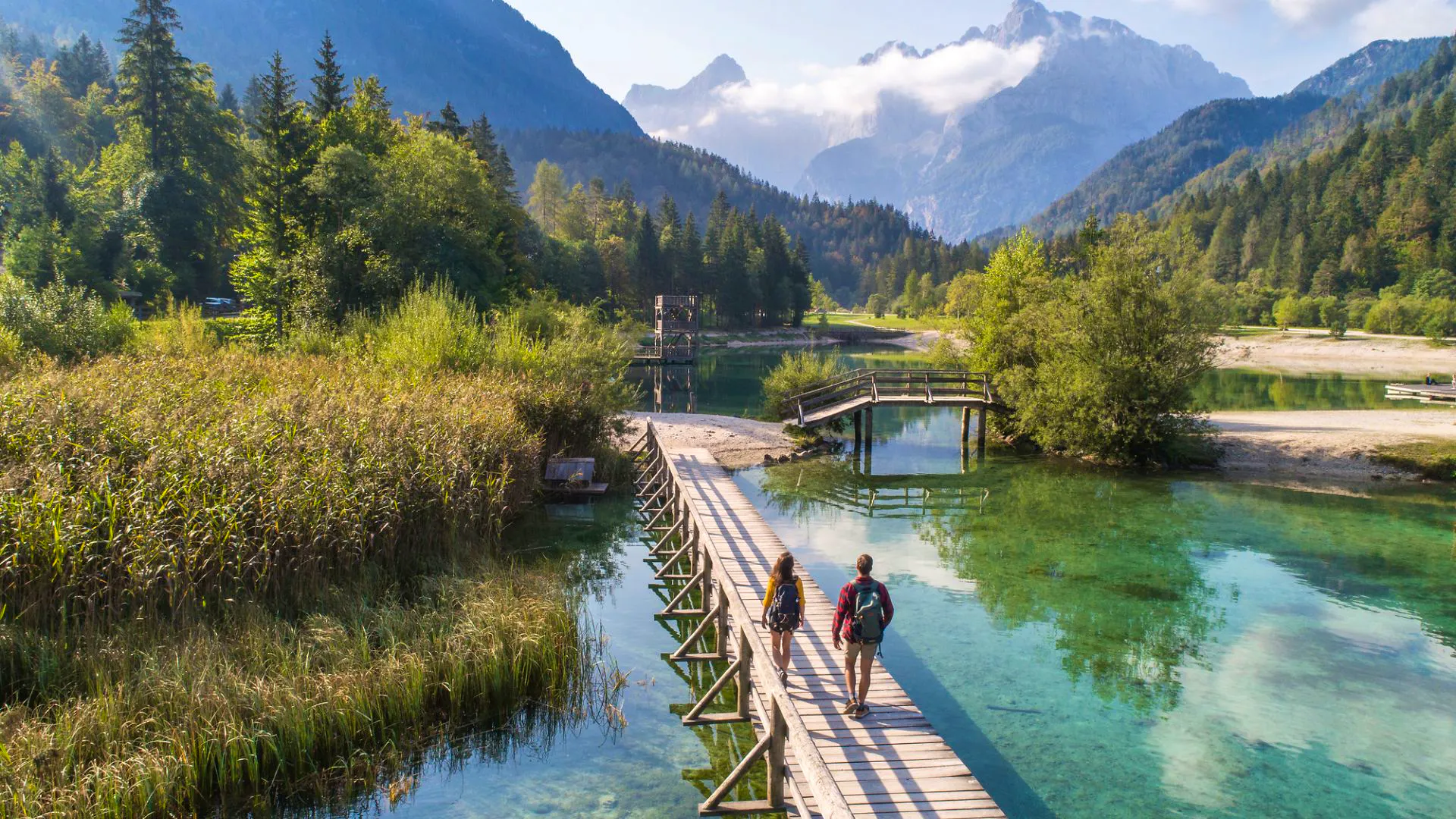 Two walkers on a wooden boardwalk over a clear green lake with Slovenian mountains behind
