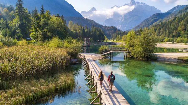Two walkers on a wooden boardwalk over a clear green lake with Slovenian mountains behind